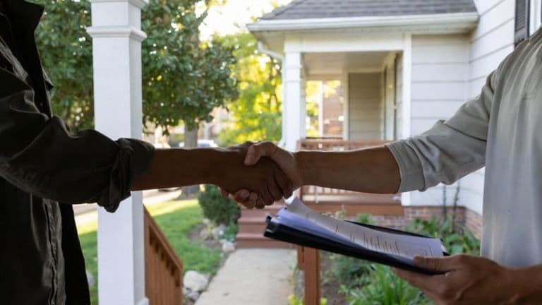 Two people shake hands outside a home while one holds documents on a clipboard.