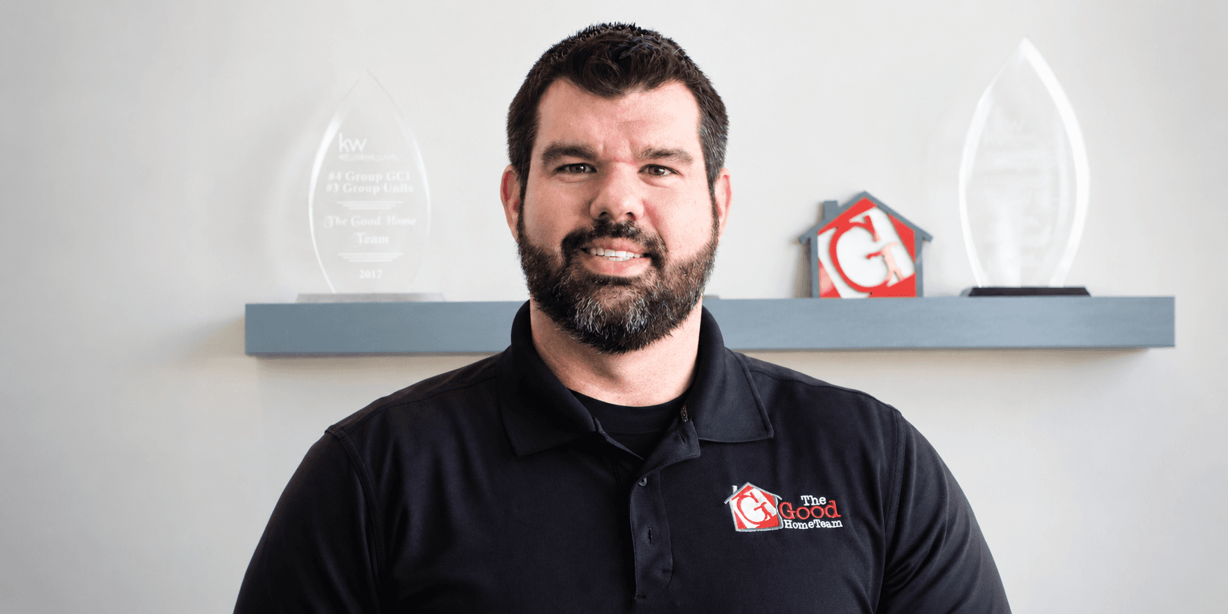 Nick Good wearing a black The Good Home Team polo with company awards displayed behind him.