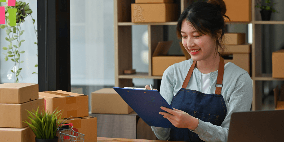 Smiling woman in an apron holds a clipboard, surrounded by cardboard boxes and a laptop