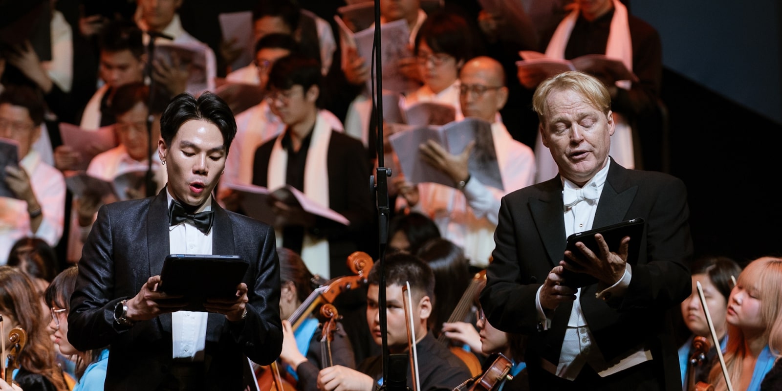 A male and female performer sing during a cultural event, with a choir and orchestra performing in the background.
