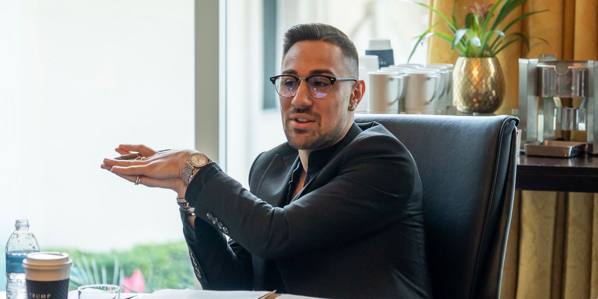 A man in glasses and a black suit gestures expressively while seated at a conference table. Coffee cups and a plant decorate the background, conveying a professional setting.