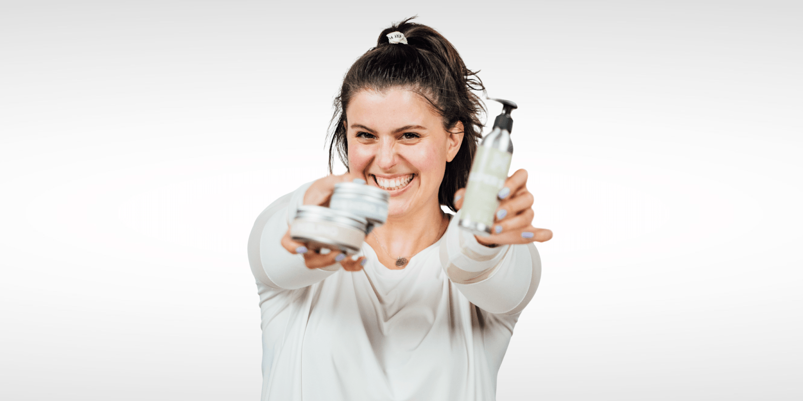 "A smiling woman in a white shirt holding skincare products in both hands, with a joyful expression, promoting wellness and beauty."