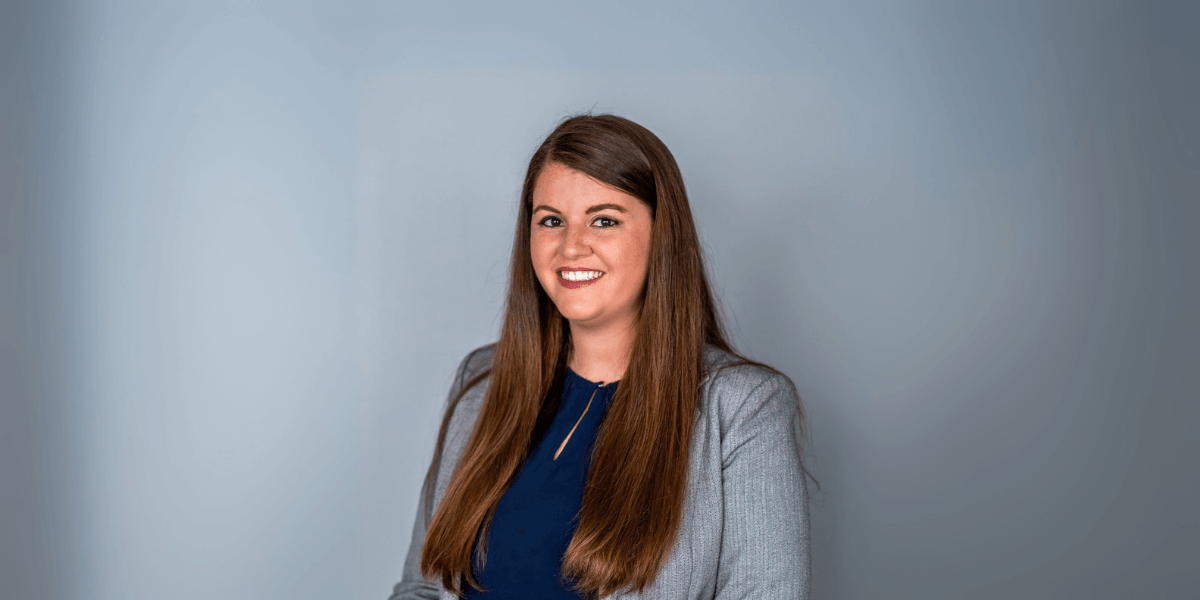 "Professional headshot of a woman smiling in a business suit, with long brown hair and a blue blouse, standing against a neutral background."
