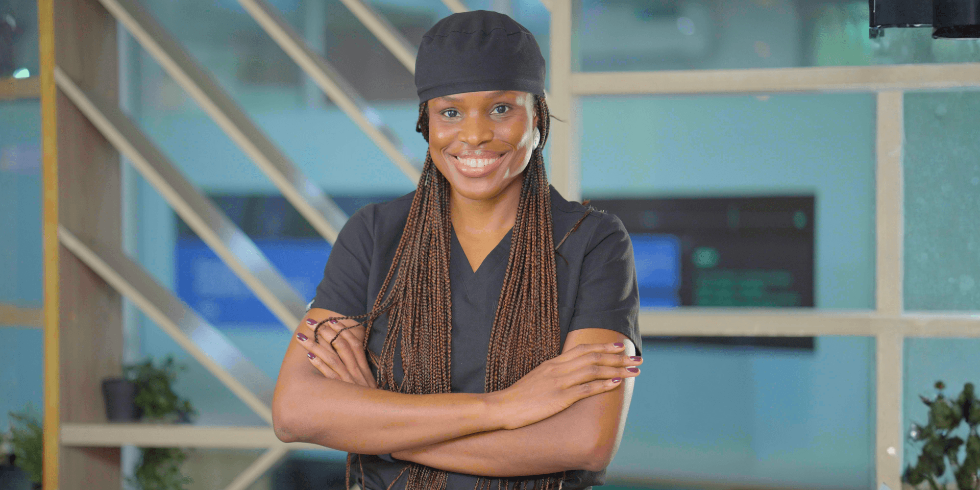 Smiling female physician in black scrubs with braided hair, standing confidently with arms crossed indoors.