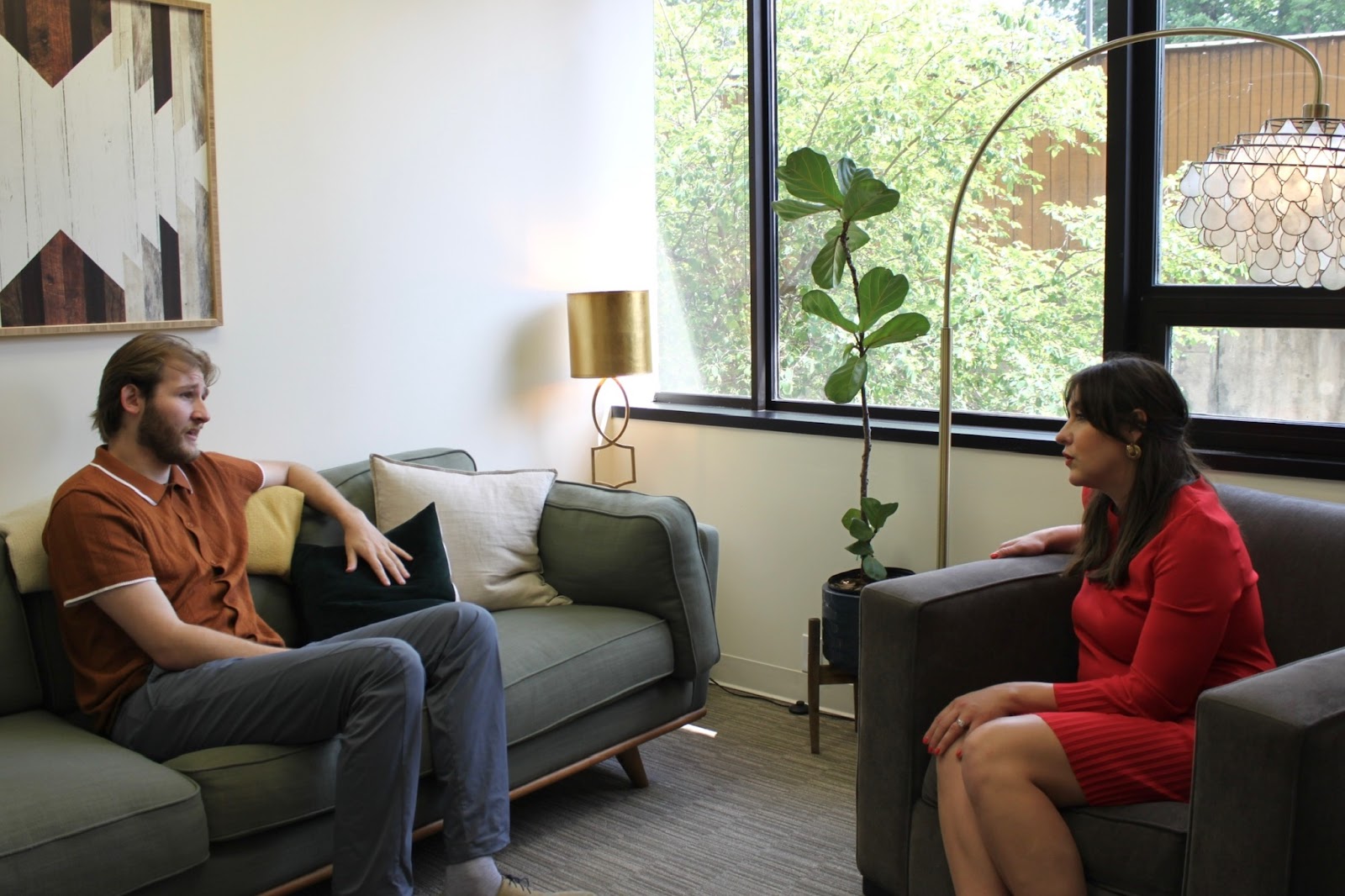 A man and woman engaged in a conversation while sitting in a cozy, well-lit living room. The man is sitting on a green couch, wearing an orange shirt and gray pants, while the woman sits in a gray armchair, dressed in a red dress. A large window lets in natural light, with a plant and lamp visible in the background.