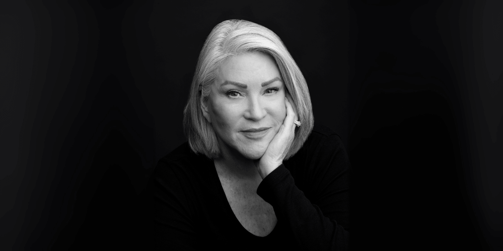 Black and white portrait of a confident woman with short silver hair, resting her chin on her hand, smiling gently against a dark background.