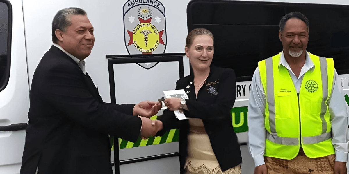 Group of three people exchanging a handshake during an official handover ceremony in front of an ambulance. The individuals include a man in a suit, a woman in formal attire holding documents, and a man in a yellow safety vest, with the Kingdom of Tonga Ambulance Service logo visible in the background.
