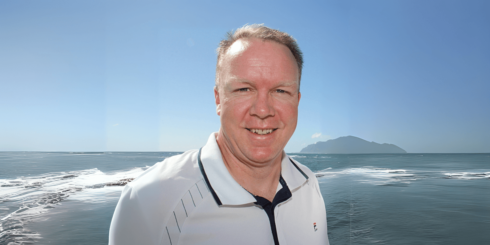 Man in a white polo shirt smiling in front of a calm ocean with a distant island under a clear blue sky.