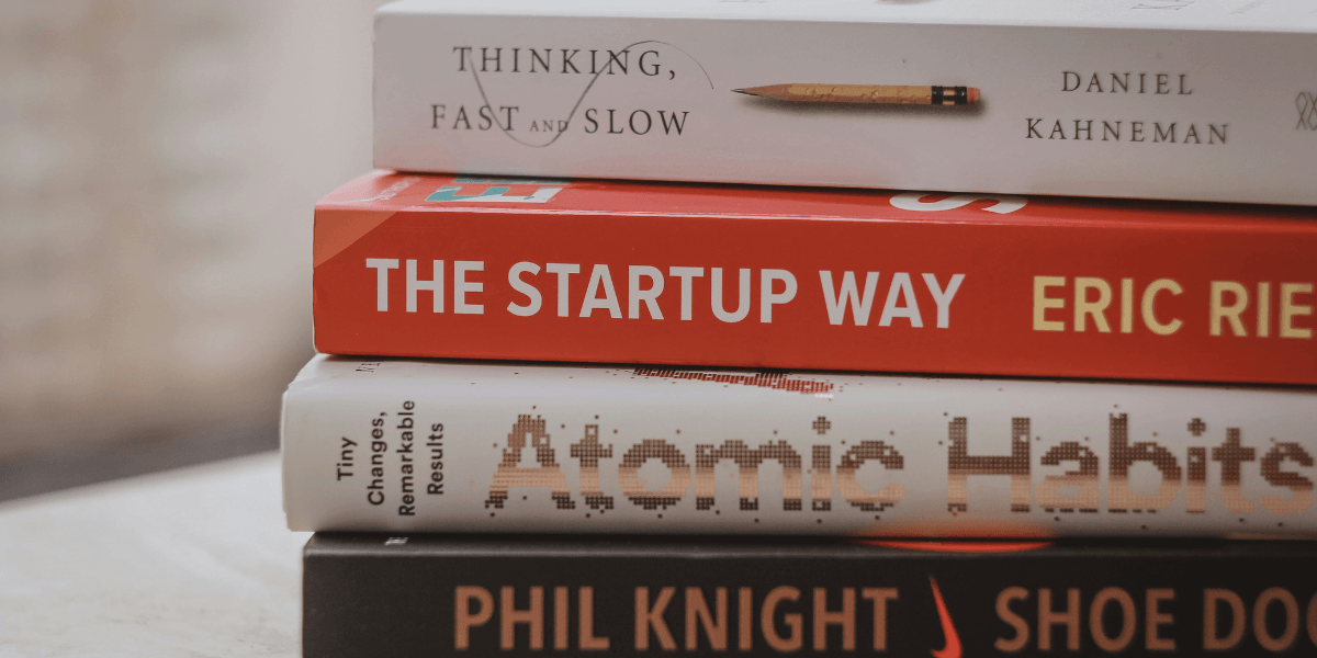 Stacked books including 'Thinking, Fast and Slow' by Daniel Kahneman, 'The Startup Way' by Eric Ries, 'Atomic Habits' by James Clear, and 'Shoe Dog' by Phil Knight, with a close-up view of their spines.