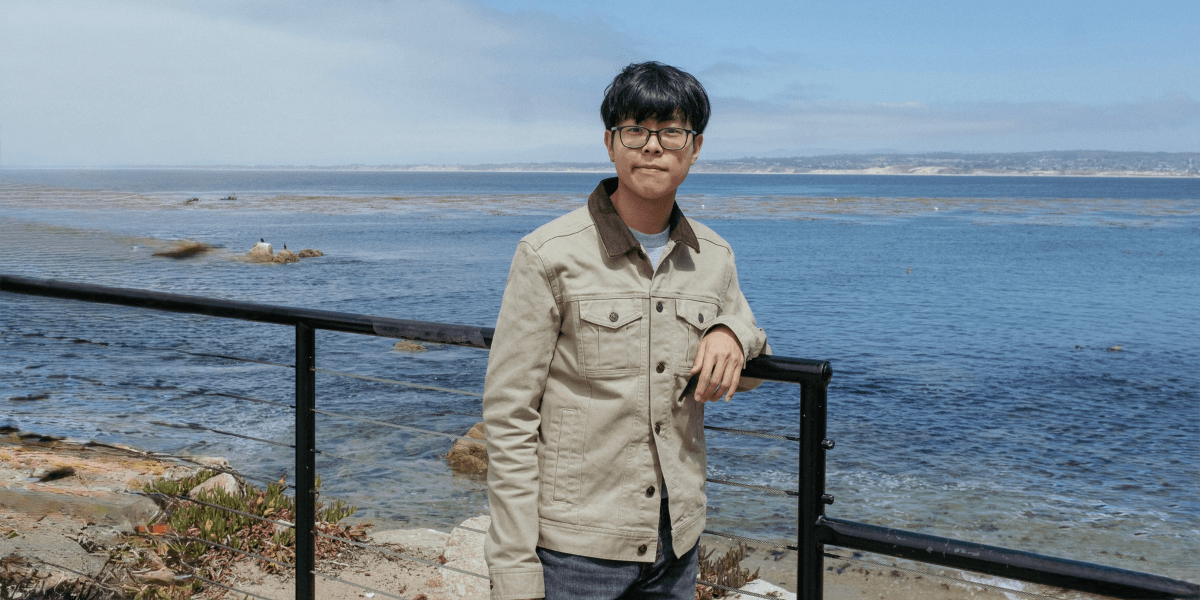 A man wearing a beige jacket and glasses standing by a fence with a scenic ocean view in the background on a sunny day.