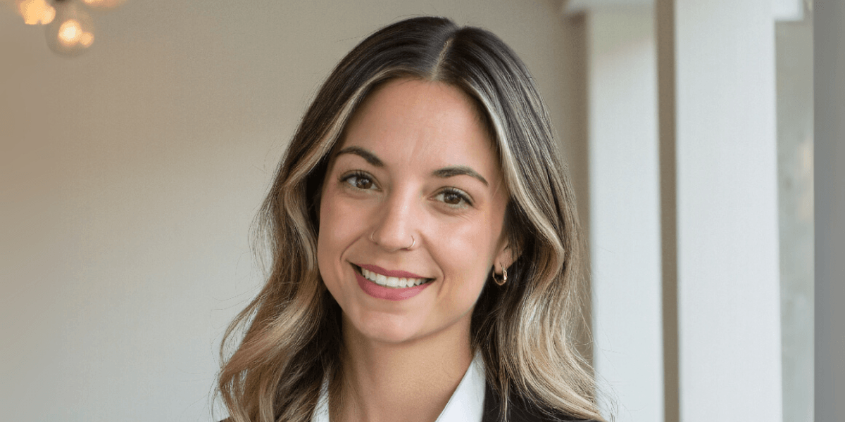 "Portrait of a smiling young woman with medium-length hair wearing a white shirt and black blazer. She has a nose piercing and gold hoop earrings, standing in a bright and modern interior space with soft lighting."