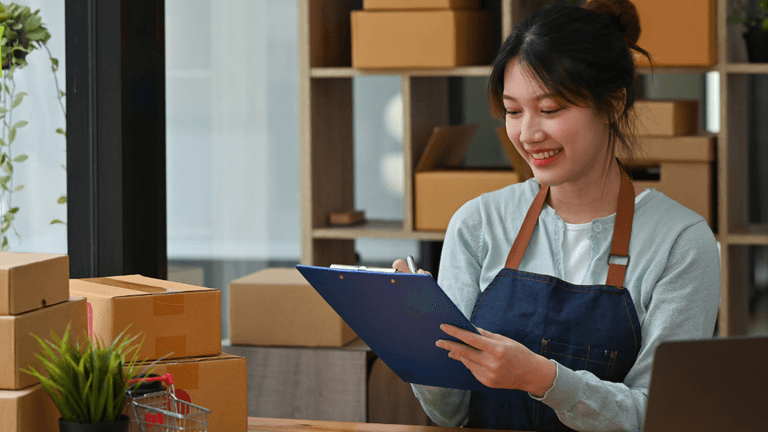 Smiling woman in an apron holds a clipboard, surrounded by cardboard boxes and a laptop