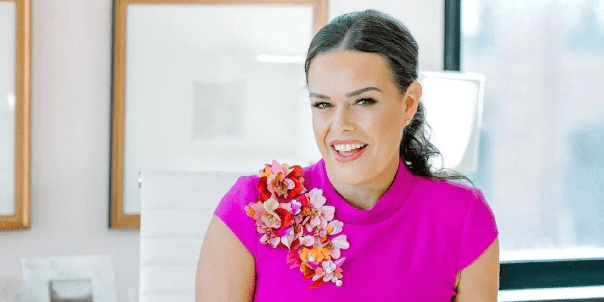 Woman in a bright pink top with floral embellishments smiles confidently in an office setting