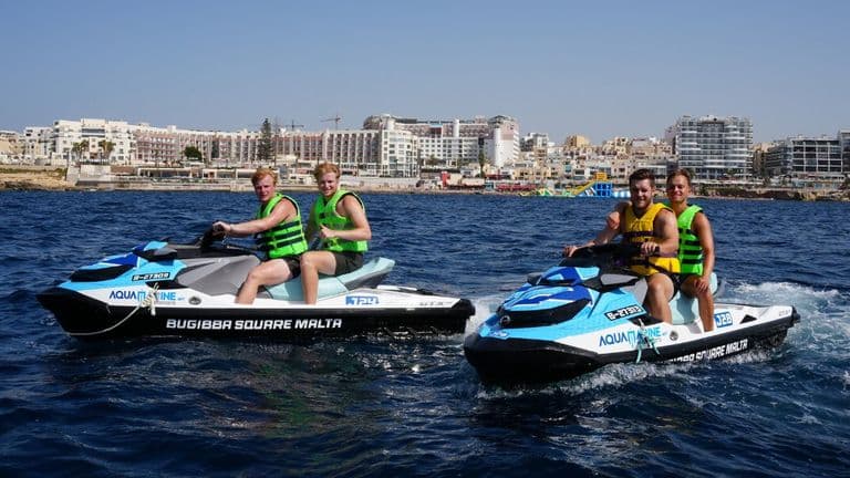 Two Aquamarine jet skis carry four riders across the water near the Bugibba waterfront.