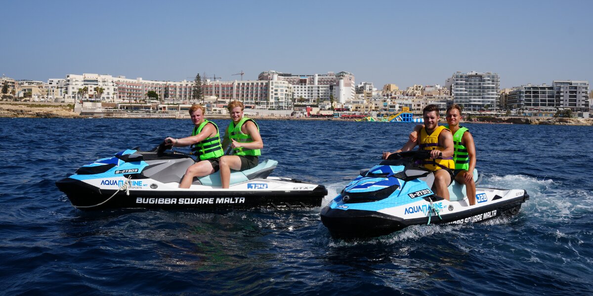 Two Aquamarine jet skis carry four riders across the water near the Bugibba waterfront.