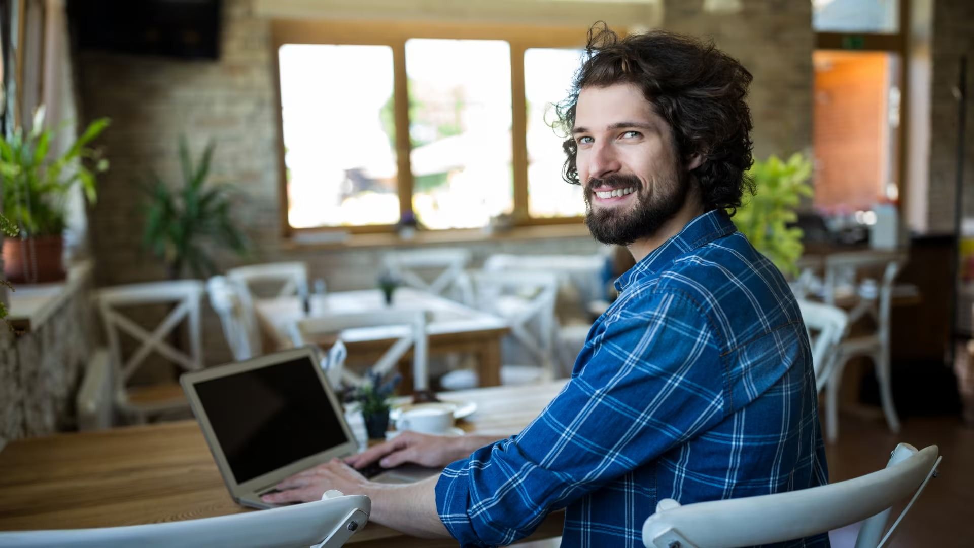 A man with curly hair and a beard smiles at the camera while using a laptop in a cozy cafe. Sunlight filters through large windows, creating a warm ambiance.