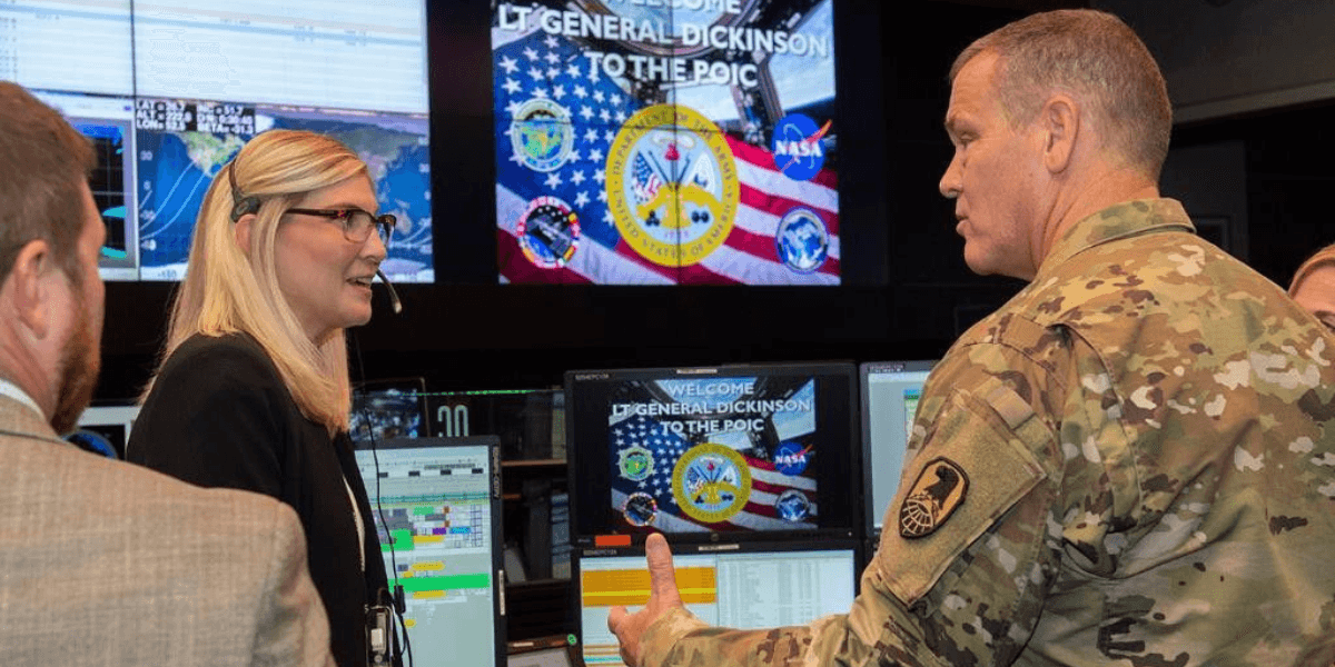 Military officer speaks with personnel inside a command center surrounded by computer monitors and U.S. military displays.